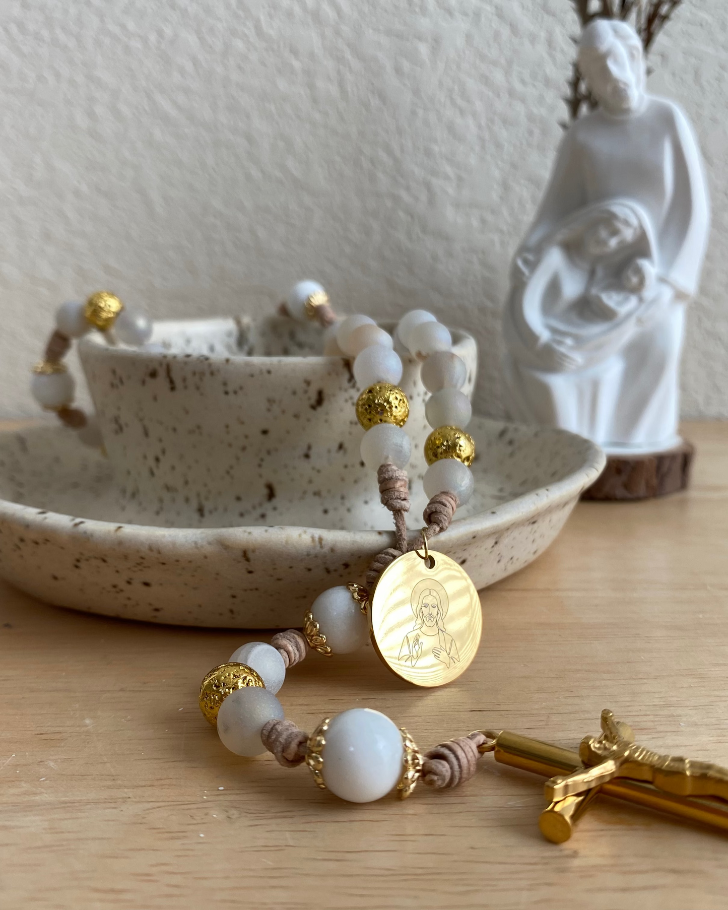 Close-up of a gold Jesus medal and gold crucifix draped over a ceramic dish with a Holy Family statue behind it.
