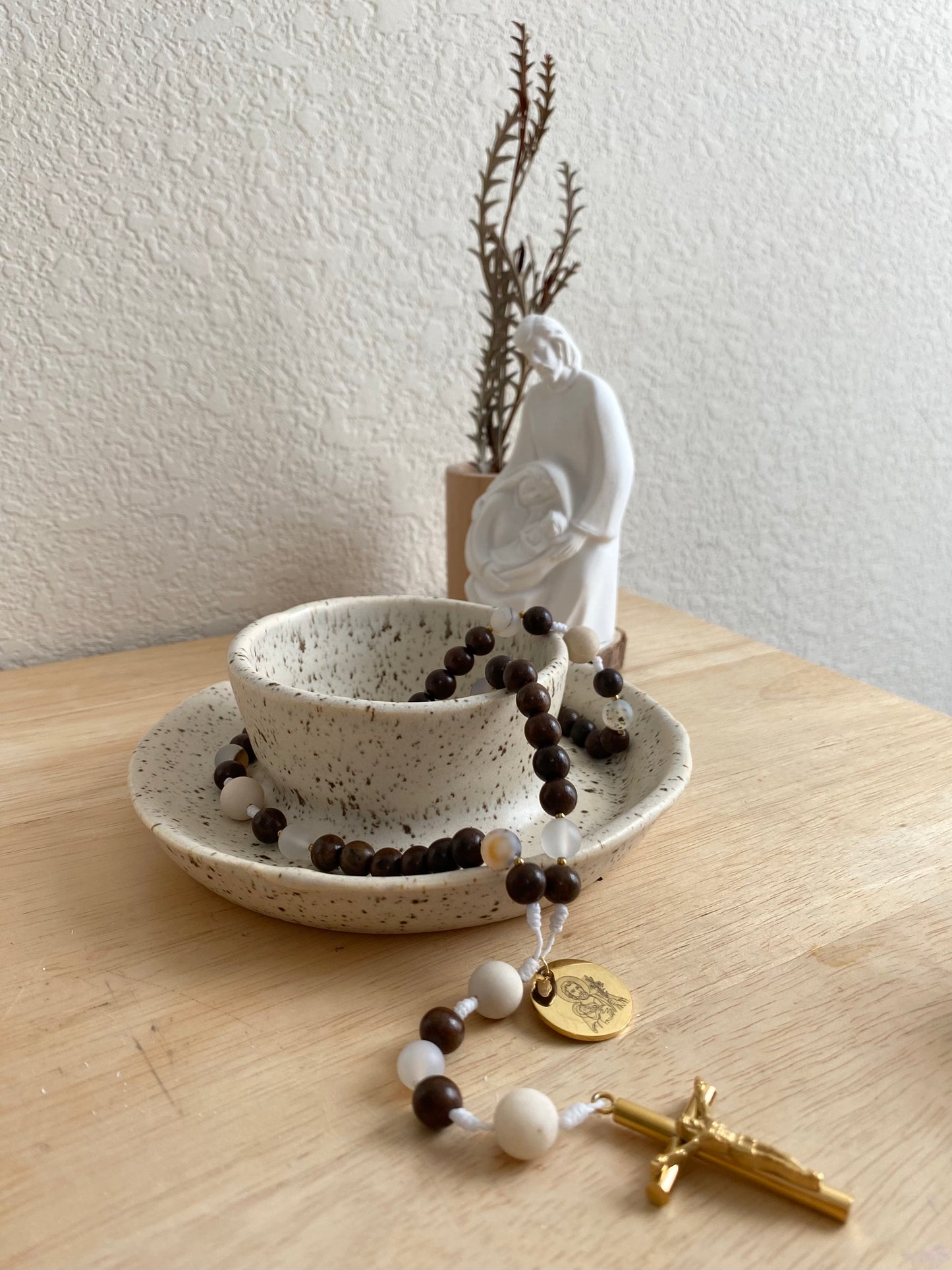 A minimal aesthetic St. Joseph rosary draped elegantly over a speckled ceramic bowl with a white Holy Family statue in a prayerful home decor setting.