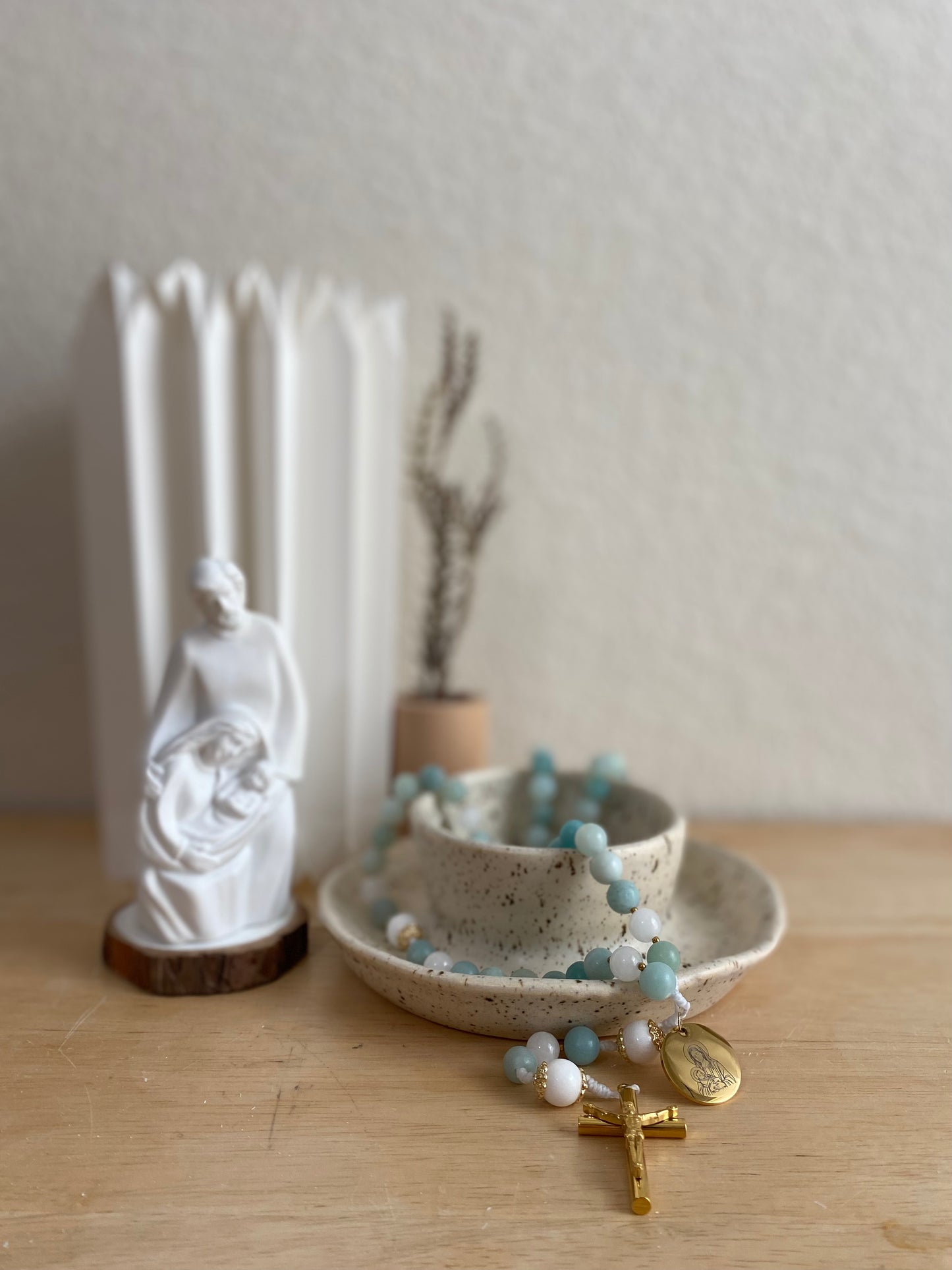 The Mary Catholic rosary draped over a speckled ceramic bowl next to a white Holy Family statue in a prayerful home setting.