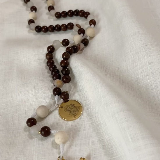 A close-up loop video showcasing the premium textures of The Joseph Catholic rosary, highlighting the smooth wood, detailed fossil jasper, and a gleaming gold Saint Joseph engraved medal on a textured white linen background.