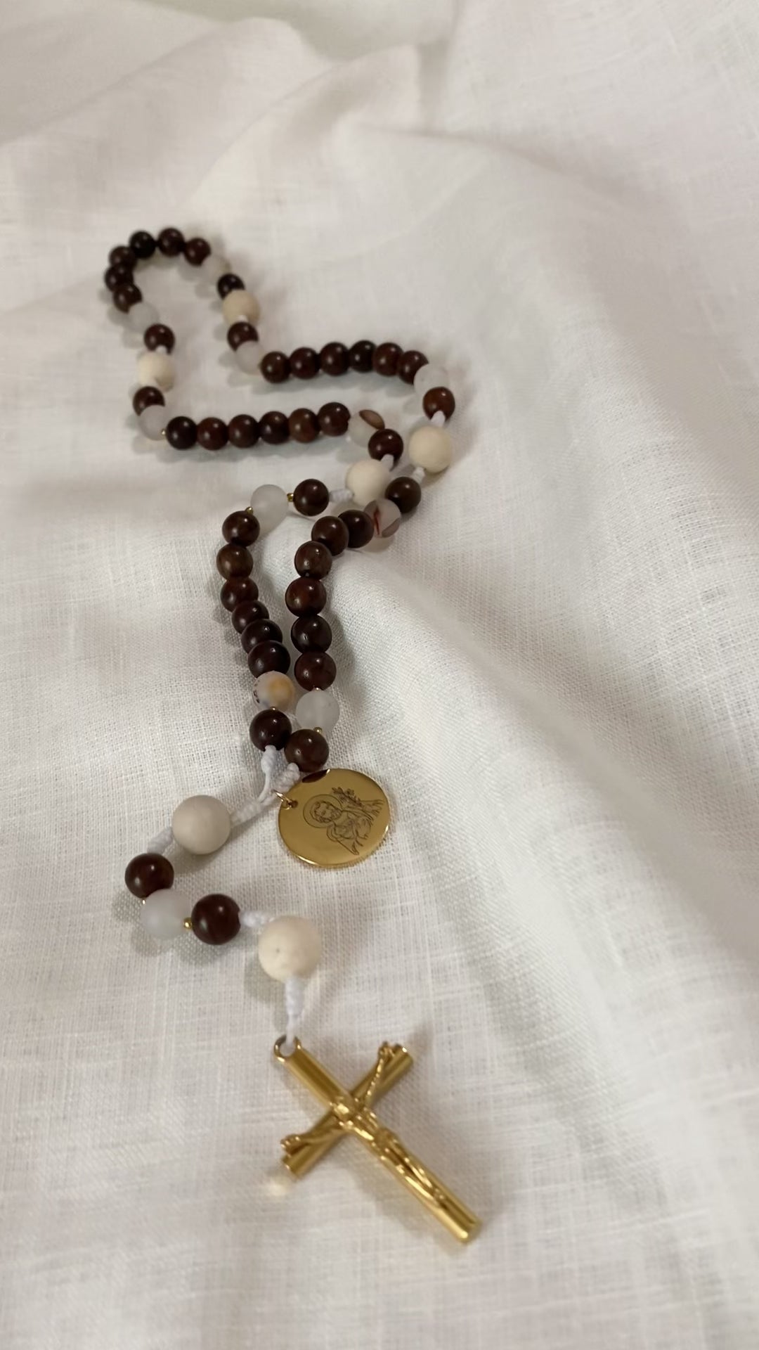 A close-up loop video showcasing the premium textures of The Joseph Catholic rosary, highlighting the smooth wood, detailed fossil jasper, and a gleaming gold Saint Joseph engraved medal on a textured white linen background.