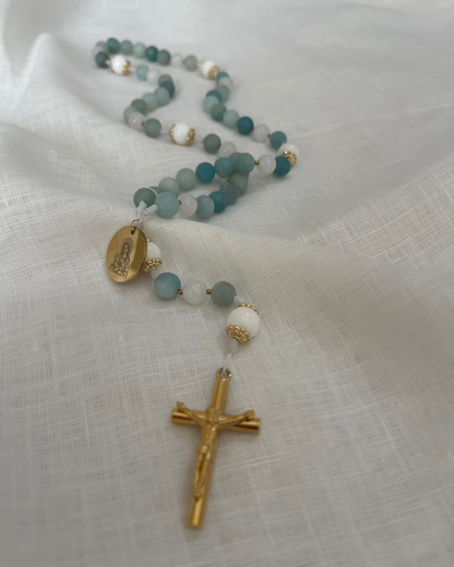 The Mary Catholic rosary made with light blue amazonite and white jade beads laid across textured white linen cloth.